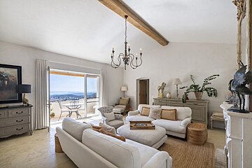 Bright, elegant living room with white sofas, a rustic wood beam, a chandelier, and glass doors opening to a scenic balcony view.