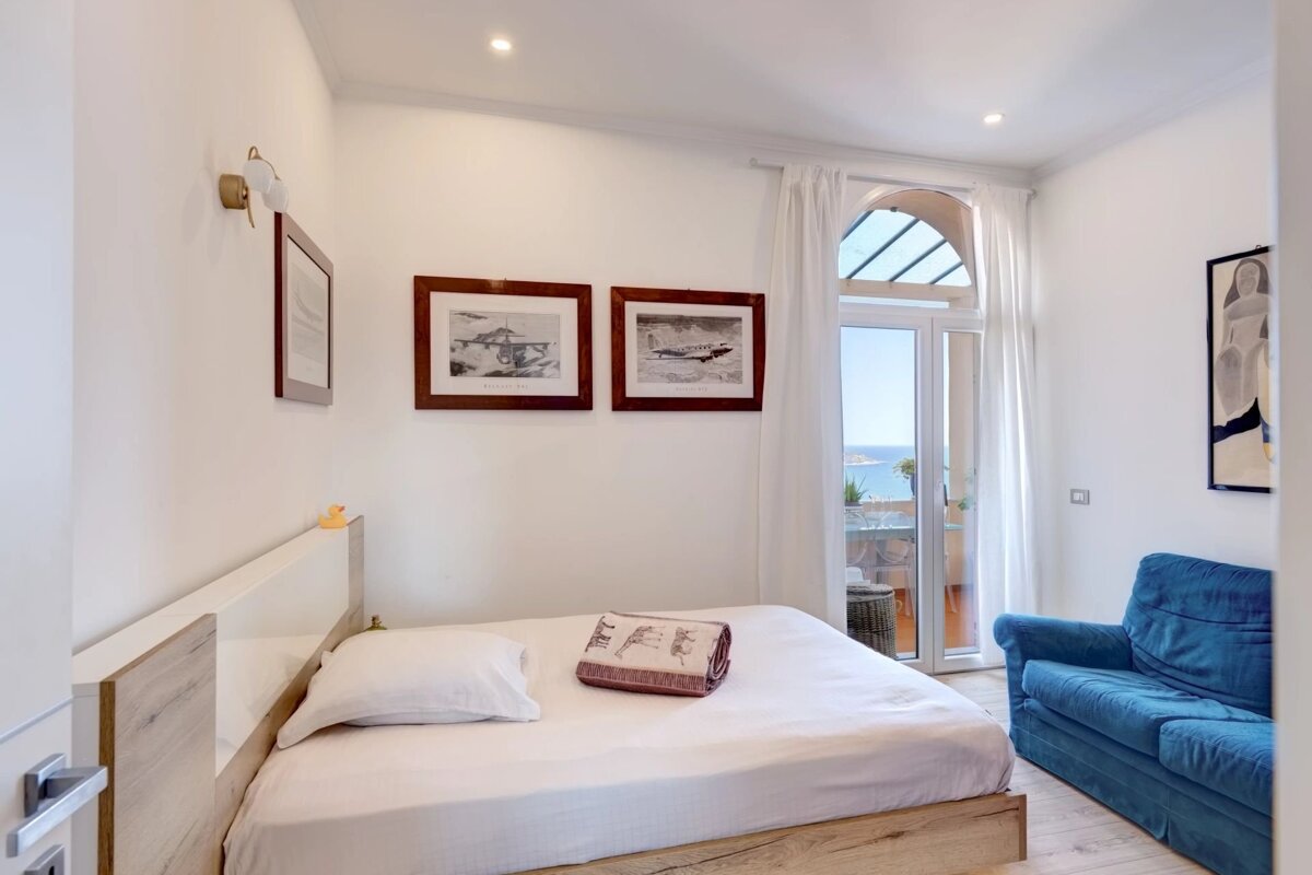 Bright bedroom with a double bed, pictures of planes, a blue sofa, and glass doors opening to a balcony with sea views.