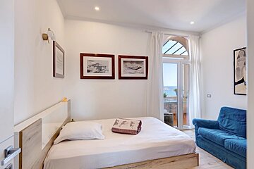 Bright bedroom with a double bed, pictures of planes, a blue sofa, and glass doors opening to a balcony with sea views.
