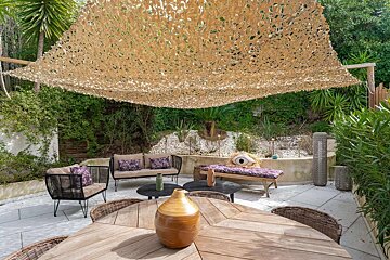 An inviting outdoor patio with a camouflage shade net, comfortable seating, tables, and a chaise lounge, set against a lush green garden.