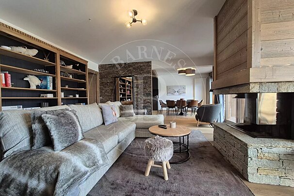 A cozy, modern chalet living room with a large grey sofa, stone fireplace, wooden bookshelves, and an open dining area in the background.