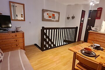 A cozy living/dining area with light wood floors, a set dining table, TV on a dresser, and a dark wood staircase railing.
