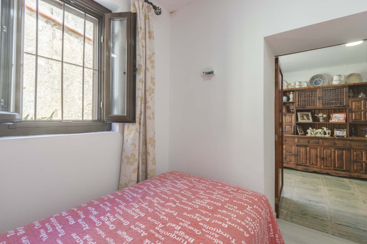 A small room features an open window with security bars, patterned curtains, and a red word-patterned bedspread. A doorway opens to another room with a large wooden cabinet.