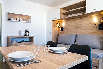 A cozy studio apartment with a dining table set for two, a gray sofa, and a kitchenette area with a kettle and coffee maker. Wooden accents and wall sconces.