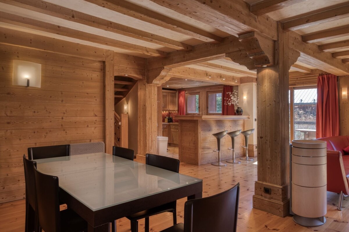 A rustic wooden chalet interior with a glass-topped dining table, exposed ceiling beams, and a bar with stools in the background.
