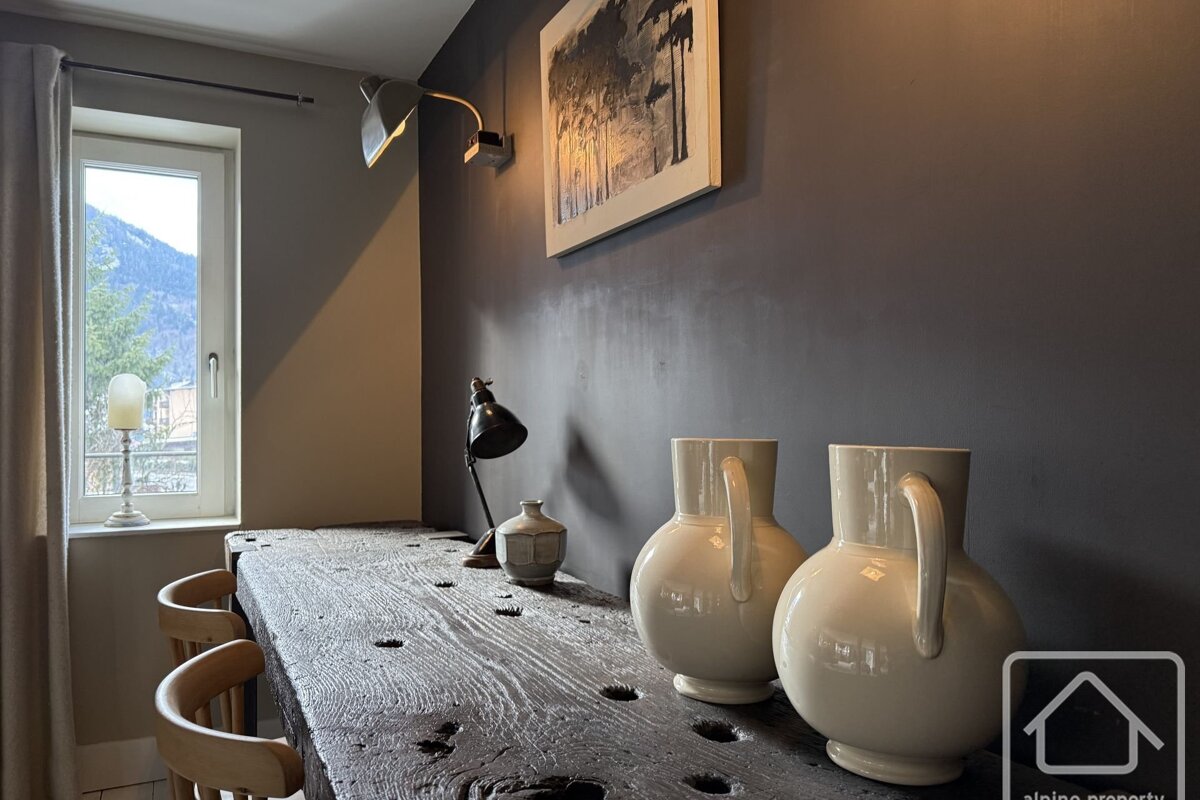 A cozy room features a rustic wooden table, modern lamps, and decorative vases. A window offers a mountain view, complemented by dark walls and framed art.