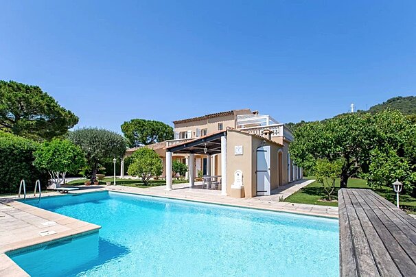 Beautiful Mediterranean villa with a large blue swimming pool, surrounded by lush green trees under a clear sunny sky.