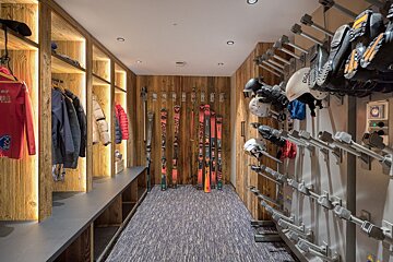 A modern ski room with warm wooden walls, illuminated lockers for jackets, skis displayed, and practical drying racks for boots and helmets.