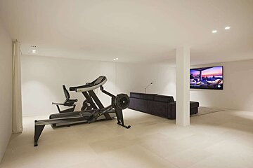 A modern, minimalist white room features a home gym (treadmill, bike, rower), a dark sofa, and a large TV displaying a vibrant sunset scene.