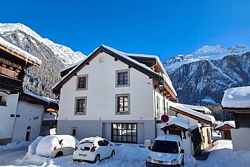 A snowy mountain village with white buildings, cars covered in snow, and majestic, snow-capped peaks under a bright blue sky.