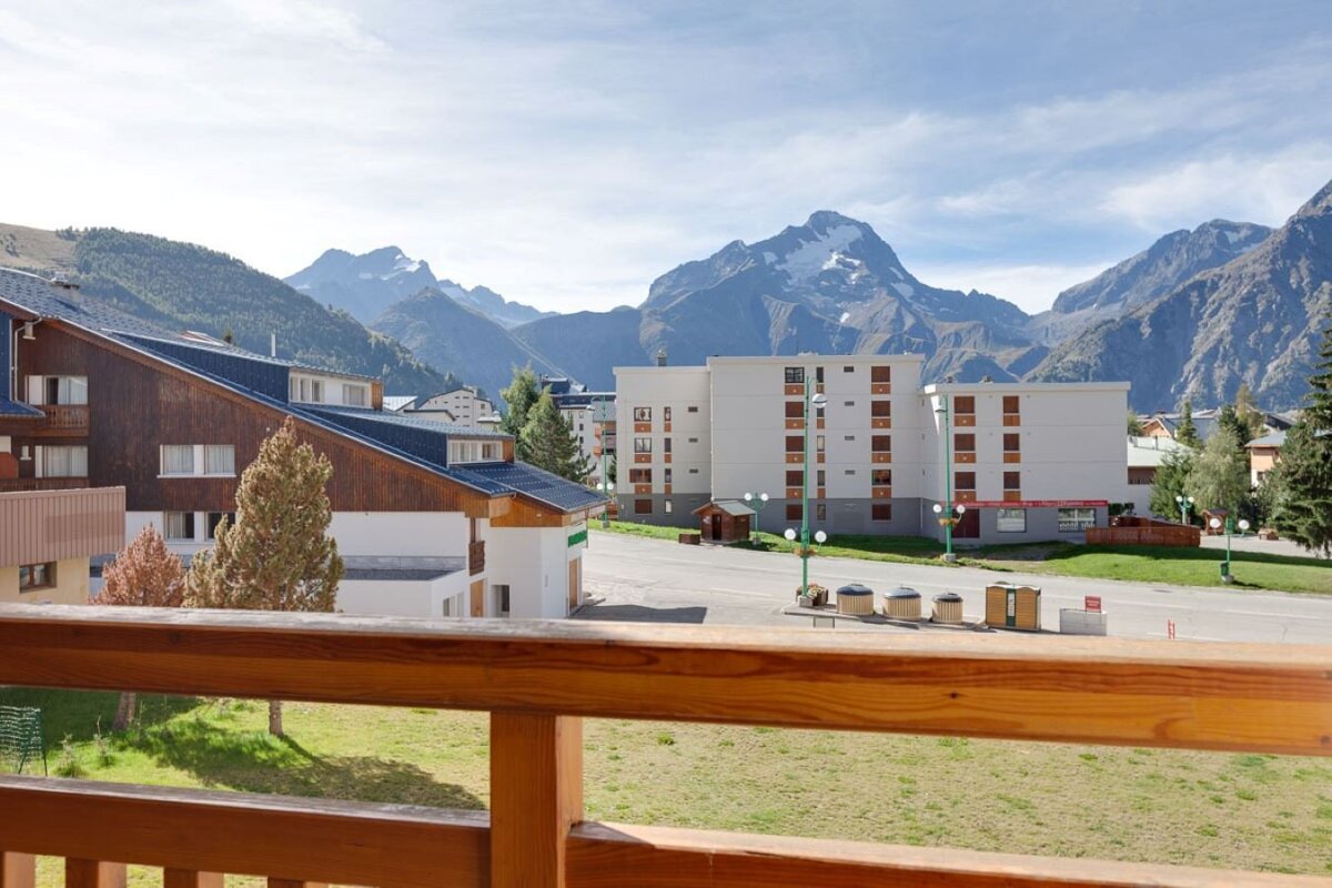 Balcony view of a mountain town. Buildings and green spaces line a road, with majestic snow-capped peaks towering in the background under blue sky.
