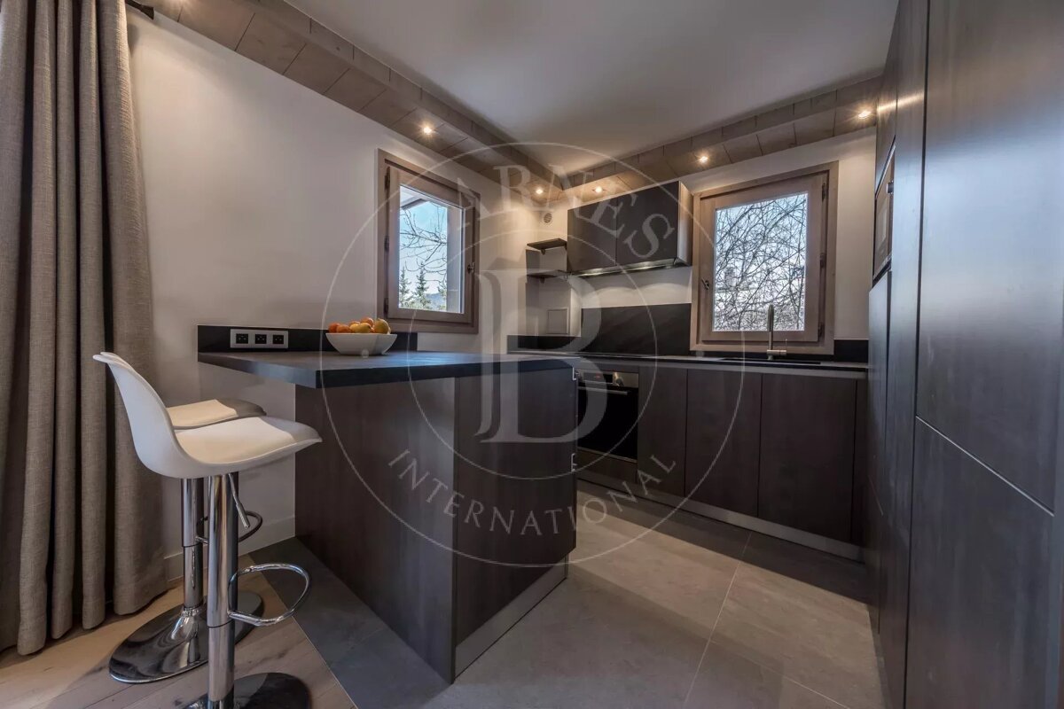 A modern kitchen featuring dark wood cabinets, a breakfast bar with a white stool, two windows, and recessed lighting.