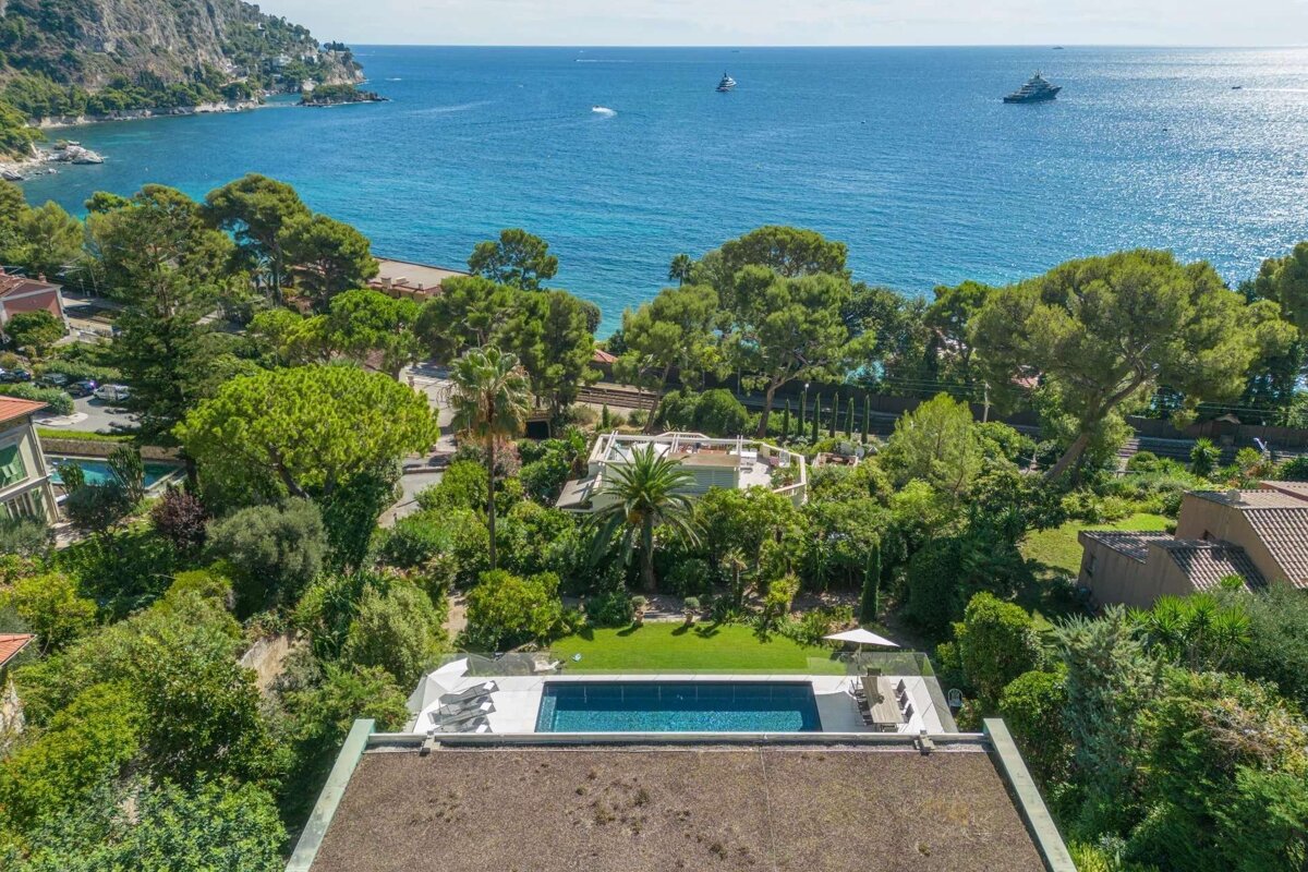 An aerial view of a luxurious coastal villa with a swimming pool, surrounded by lush greenery, overlooking the blue sea with distant boats.