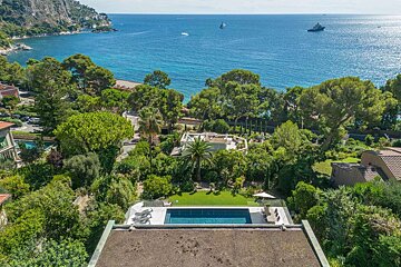 An aerial view of a luxurious coastal villa with a swimming pool, surrounded by lush greenery, overlooking the blue sea with distant boats.