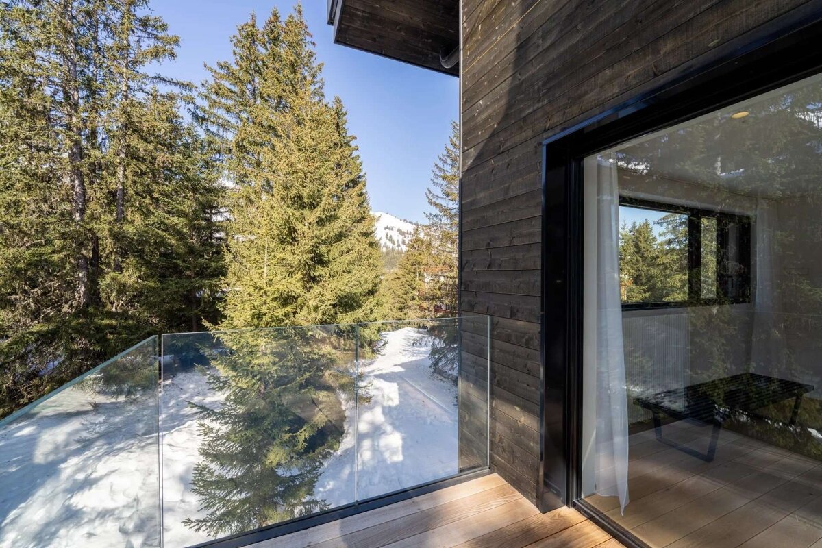 A modern wooden house balcony with a glass railing offers a sunny view of a snowy pine forest and distant mountains.