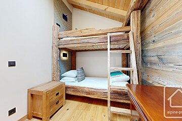 A cozy, rustic room features sturdy wooden bunk beds with a ladder, light walls, wooden floor, and exposed ceiling beams.