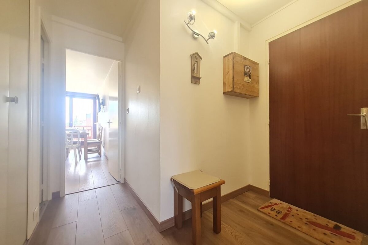 Entryway with a wooden door, stool, and small wall cabinet, leading down a hallway toward a brightly lit dining area.