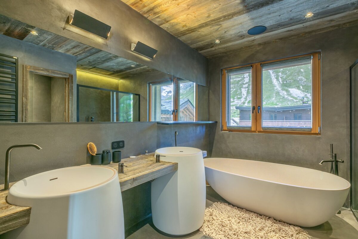 A modern, rustic bathroom featuring two pedestal sinks, a freestanding tub, and a wood ceiling. Windows offer a scenic mountain view.