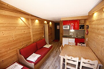 A cozy, wood-paneled studio apartment with a red sofa, wooden dining table, and compact kitchen with red cabinets. Alpine chalet style.