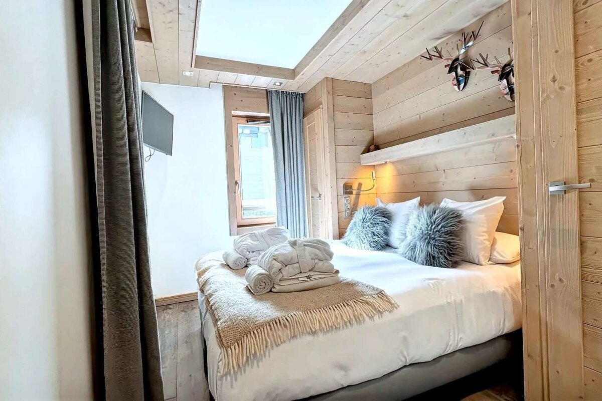 A cozy bedroom features light wood paneling, a bed with white linens, fluffy pillows, folded bathrobes, a window with curtains, and deer decor.