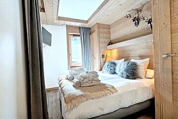 A cozy bedroom features light wood paneling, a bed with white linens, fluffy pillows, folded bathrobes, a window with curtains, and deer decor.