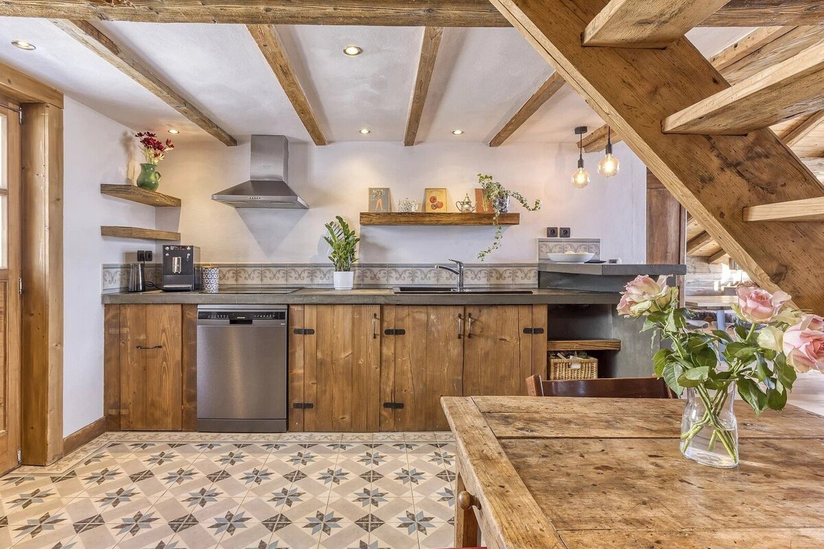 Charming rustic kitchen with wooden beams, patterned floor, wood cabinets, and a wooden dining table with flowers under a staircase.