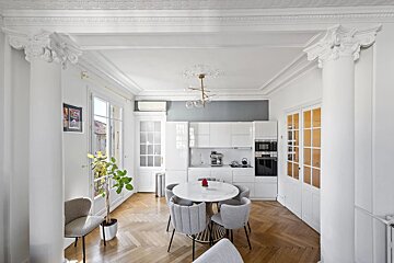 Bright, elegant room with white kitchen, round dining table, herringbone floor, ornate moldings, and a balcony view.