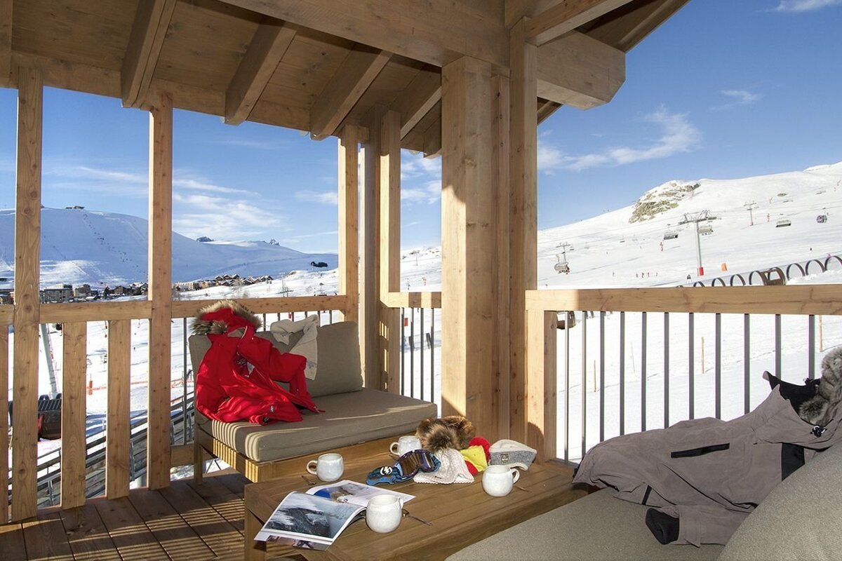 A wooden balcony offers a stunning view of a snowy ski resort with mountains and ski lifts. Mugs and ski gear sit on a table and benches.