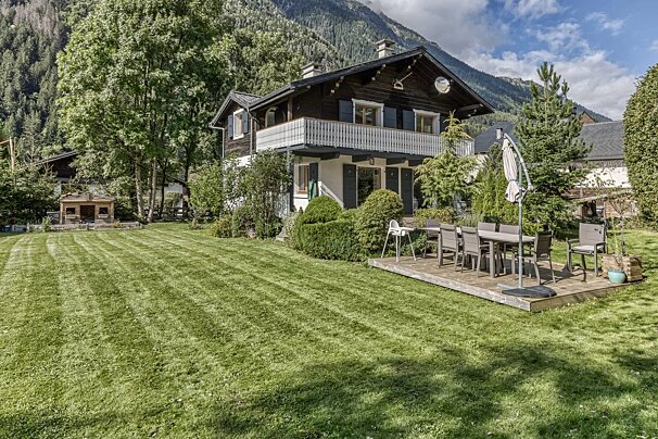 An Alpine-style house with dark wood accents and balcony, a large mowed lawn, and outdoor dining on a deck, all against a mountain backdrop.