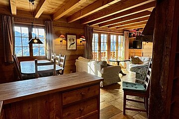 Cozy, rustic wooden cabin interior with exposed ceiling beams, dining and living areas, and mountain views through large windows.