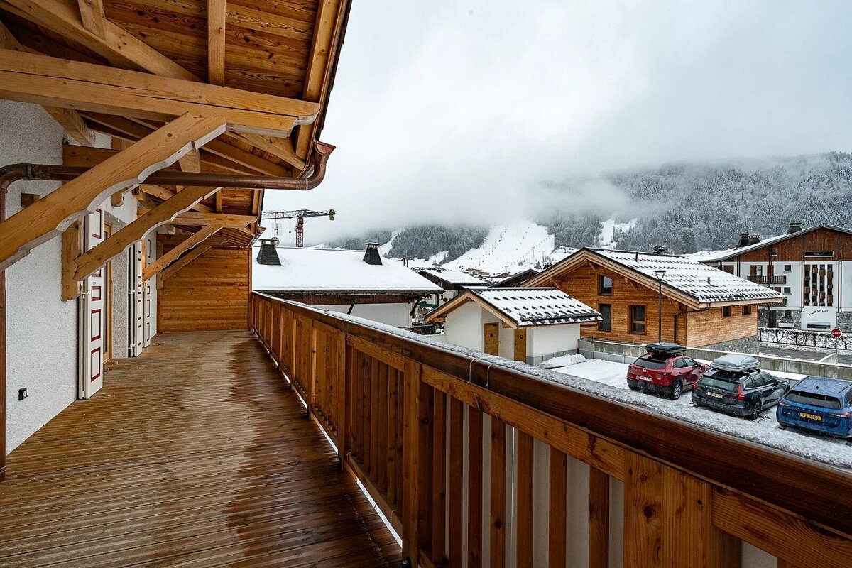 View from a wooden chalet balcony overlooking a snow-covered alpine village with misty mountains.