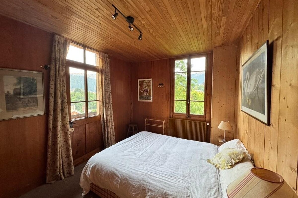 A cozy, rustic bedroom with wood-paneled walls and ceiling, a double bed with white linens, and windows showing a scenic view.