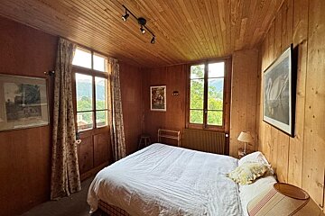 A rustic bedroom with wood-paneled walls and ceiling features a bed, two windows offering views of lush greenery, and several framed pictures.