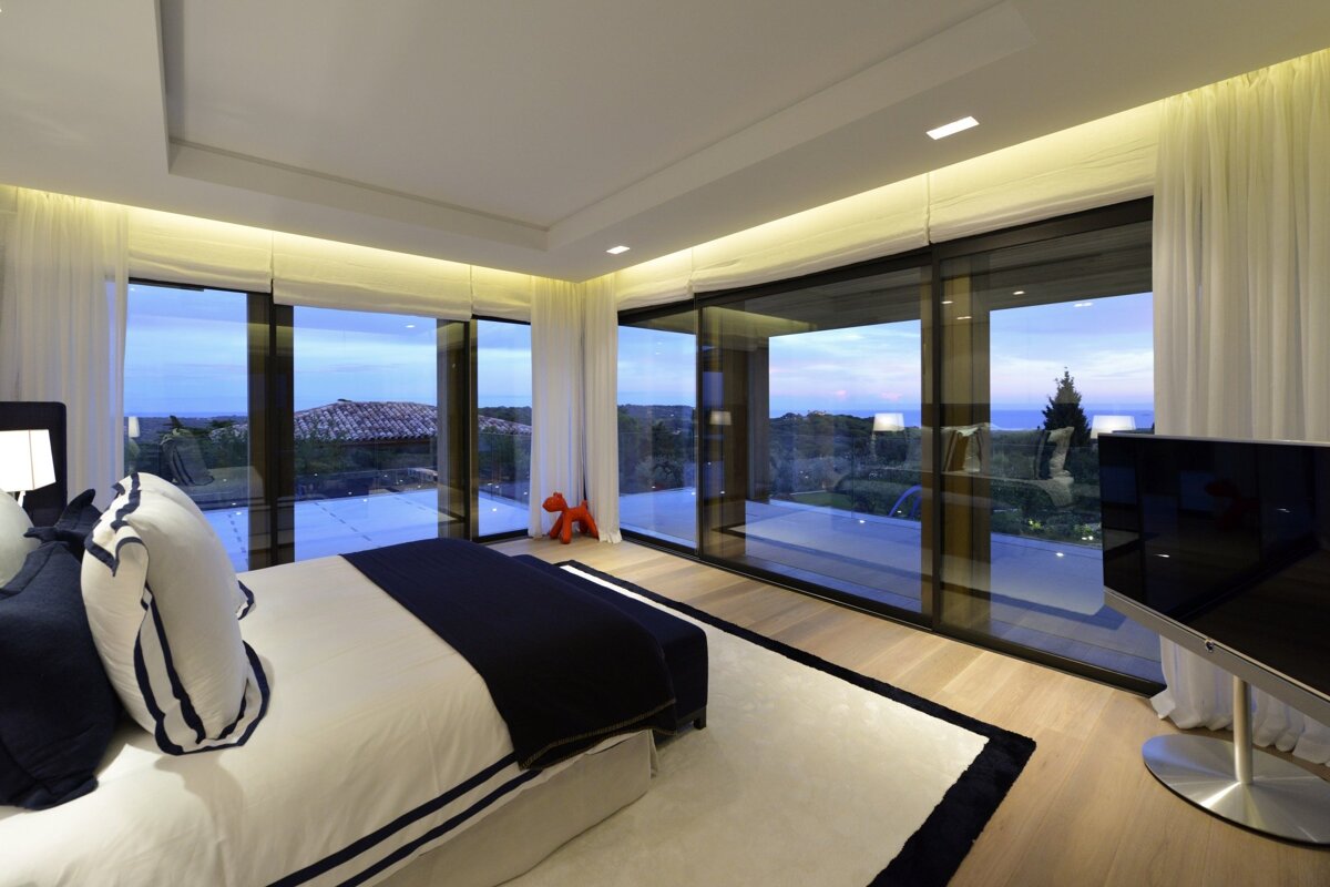 Modern bedroom with panoramic glass walls showcasing a beautiful landscape view at dusk. Navy and white decor with ambient lighting.