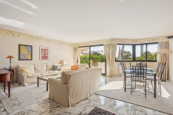 Bright open-plan living and dining room with marble flooring, cream sofas, and large windows showcasing outdoor greenery and palm trees.