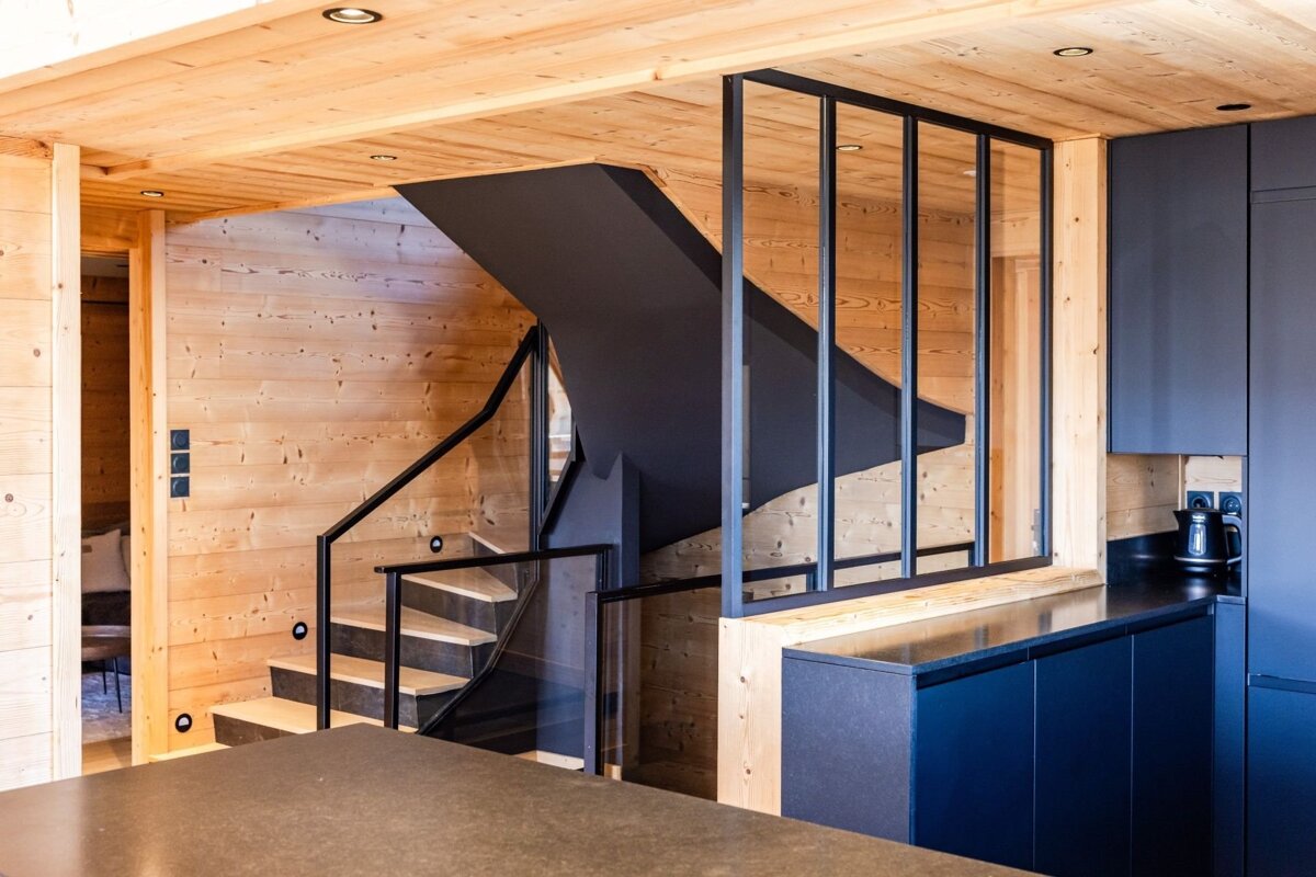 Modern wooden interior featuring a black staircase, dark blue kitchen cabinets, and a black-framed glass partition.