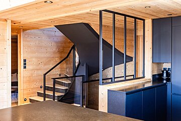 Modern wooden interior featuring a black staircase, dark blue kitchen cabinets, and a black-framed glass partition.