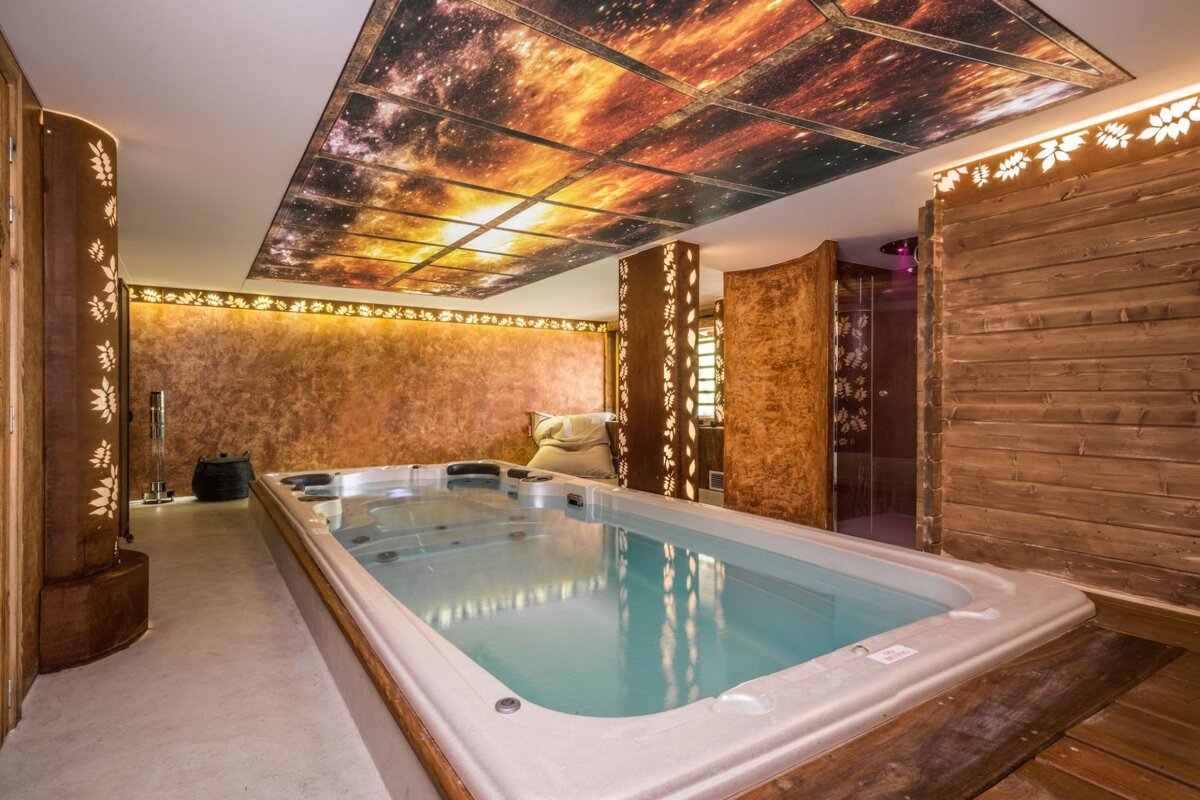 Luxurious indoor spa with a long hot tub, a glowing cosmic-themed ceiling, and warm, textured walls with decorative carved panels and lighting.