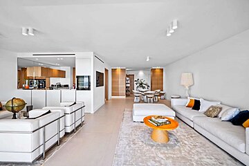A spacious, modern open-concept living room and kitchen. White decor, wood accents, and a bright orange coffee table create a sleek look.