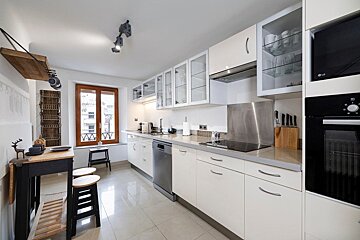 A bright, modern kitchen featuring white cabinets, stainless steel appliances, a small wooden dining table with stools, and a window.