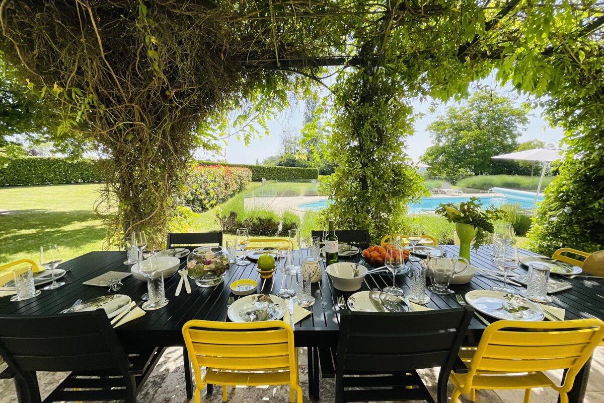 An outdoor dining table shaded by a lush green pergola, with bright yellow and black chairs, overlooking a beautiful garden and swimming pool.