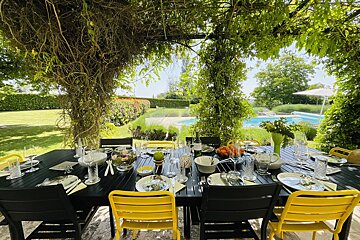 An outdoor dining table shaded by a lush green pergola, with bright yellow and black chairs, overlooking a beautiful garden and swimming pool.
