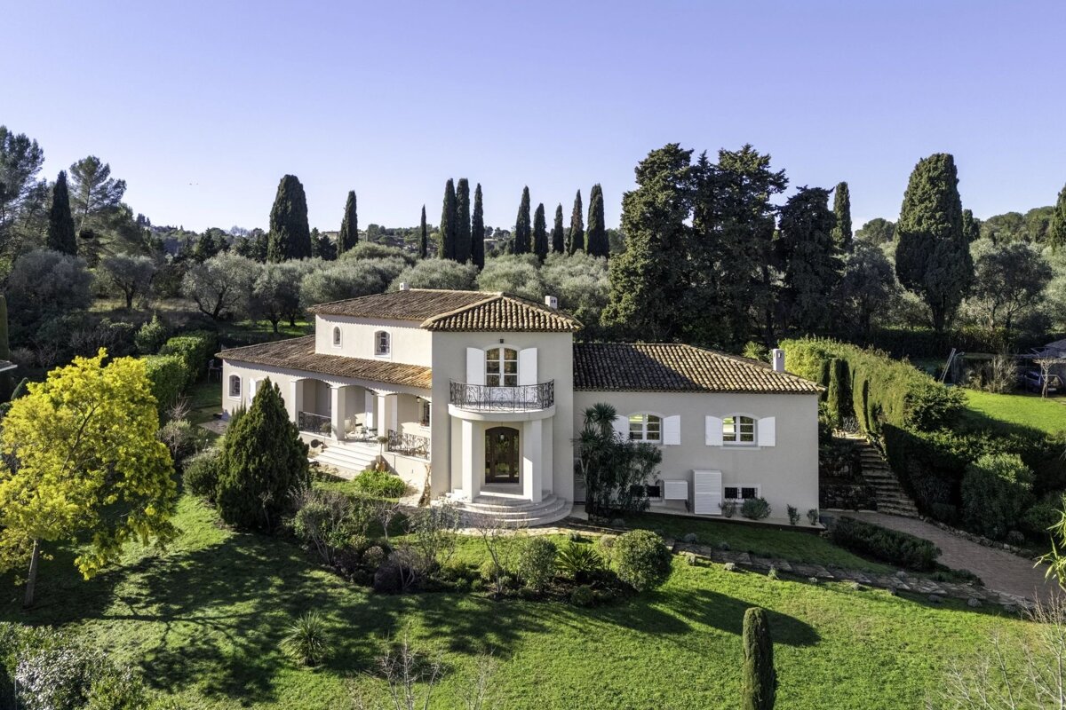 An elegant Mediterranean villa with a tiled roof, surrounded by a lush green lawn, olive groves, and tall cypress trees under a clear sky.