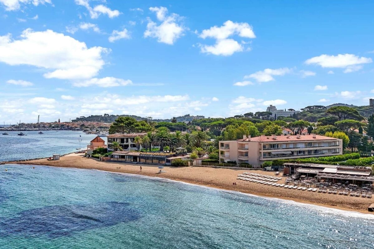 An aerial view of a sunny beach with clear turquoise water, sandy shore, beachfront resorts/buildings, and green hills, with a distant harbor town.