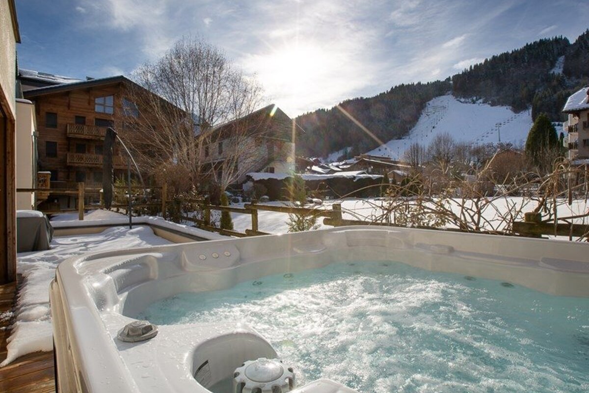 An outdoor hot tub bubbles in a snowy setting with mountain chalets and a ski slope under a bright sun.
