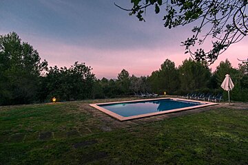 A tranquil outdoor pool at dusk under a pink-purple sky, surrounded by green foliage. Lounge chairs and a glowing lantern line the pool area.
