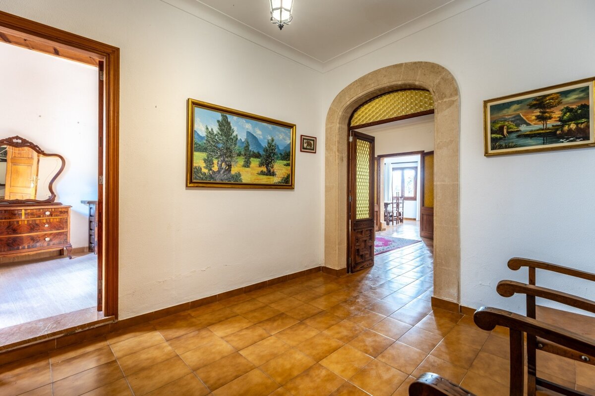 A warm-toned hallway with tiled floors, white walls, multiple paintings, an ornate stone archway, and wooden doorways to other rooms.