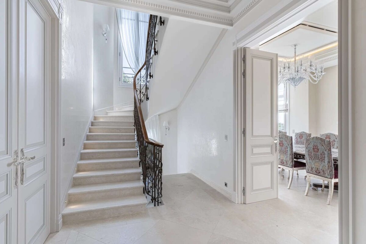 Elegant, bright foyer with a marble staircase, ornate railing, and a view into a formal dining room with a grand chandelier.