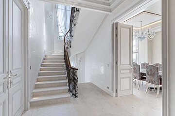 An elegant, bright white entryway features a winding staircase with a dark ornate railing. An open doorway reveals a lavish dining room.