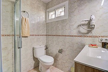 A small, tiled bathroom features a white toilet, sink, shower stall, a small window, and a wall-mounted hairdryer.